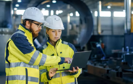 Två personer i varselkläder och vita hjälmar står i en industrimiljö och tittar på en laptop tillsammans.
